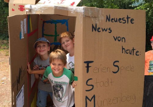 Drei Jungen schauen vergnügt aus einem angemalten Karton.